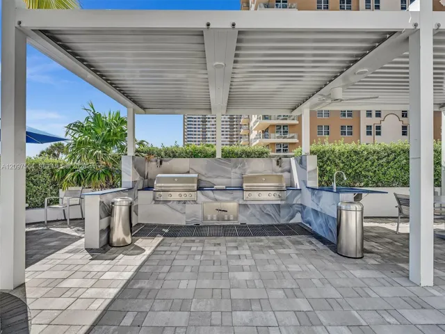 Outdoor kitchen featuring a stone-clad backsplash, dual stainless steel grills, a built-in sink, and a covered pergola structure over stone paver flooring