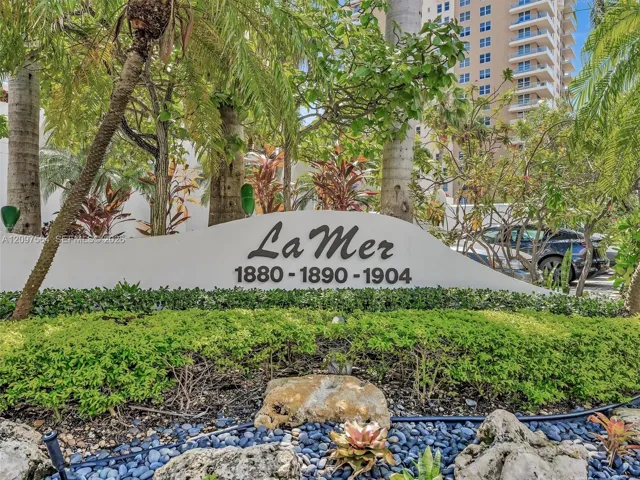 Custom signage featuring La Mer property identification, stone accent boulders, manicured shrubbery, and tropical landscaping