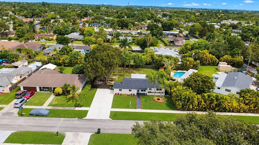 Aerial perspective of a property featuring a rectangular swimming pool, concrete driveway, lush landscaping, and mature trees on a level lot