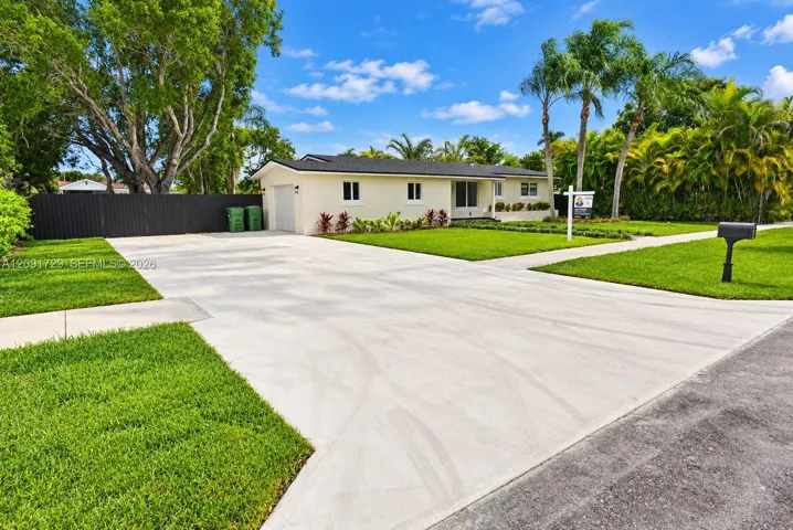 Single-story exterior featuring a light-toned stucco facade, a dark-finished roof, an expansive concrete driveway, a manicured lawn, and mature palm trees
