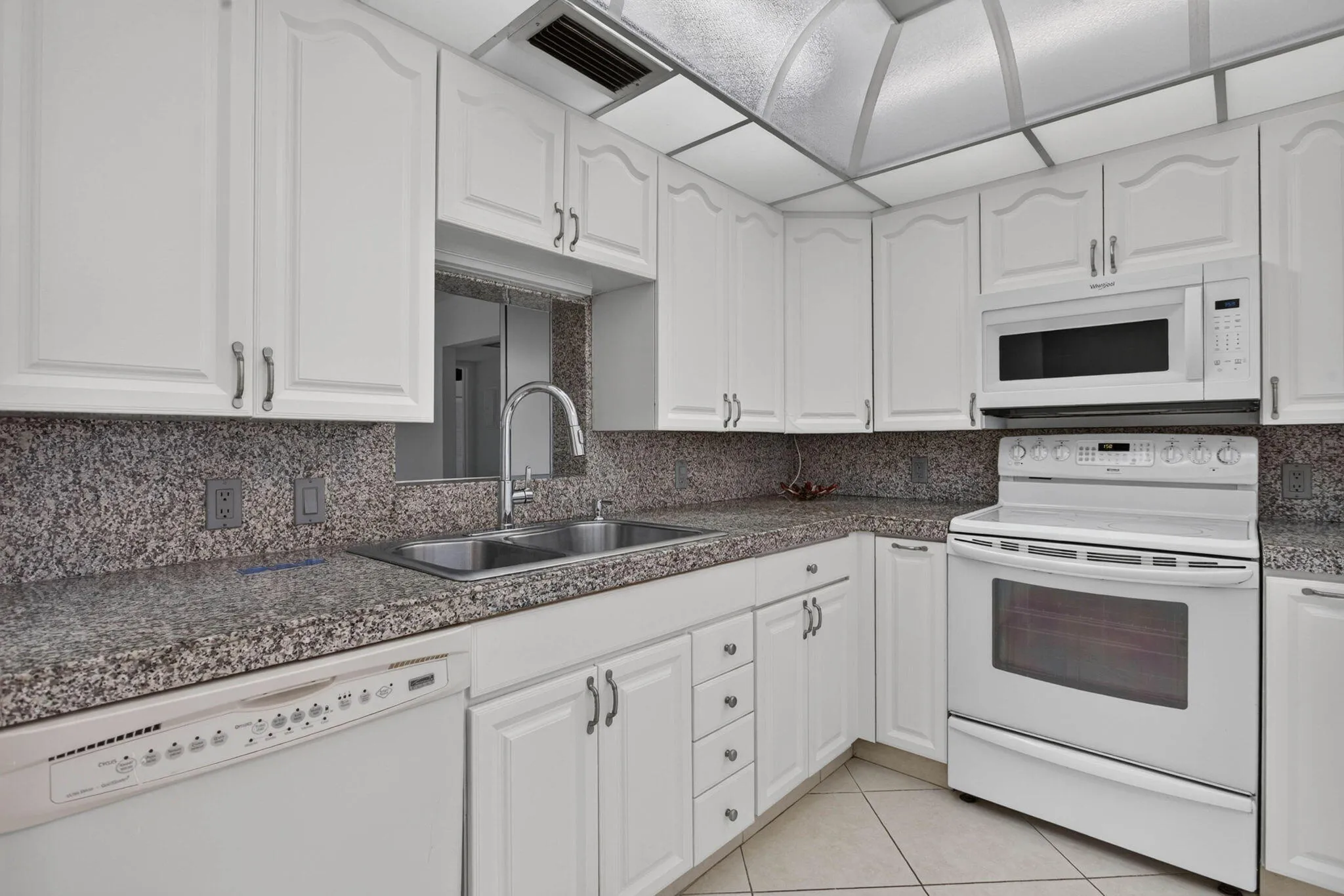 White cabinetry with silver-toned hardware, stone-finish countertops, stainless steel double basin sink with gooseneck faucet, white built-in microwave, and white range