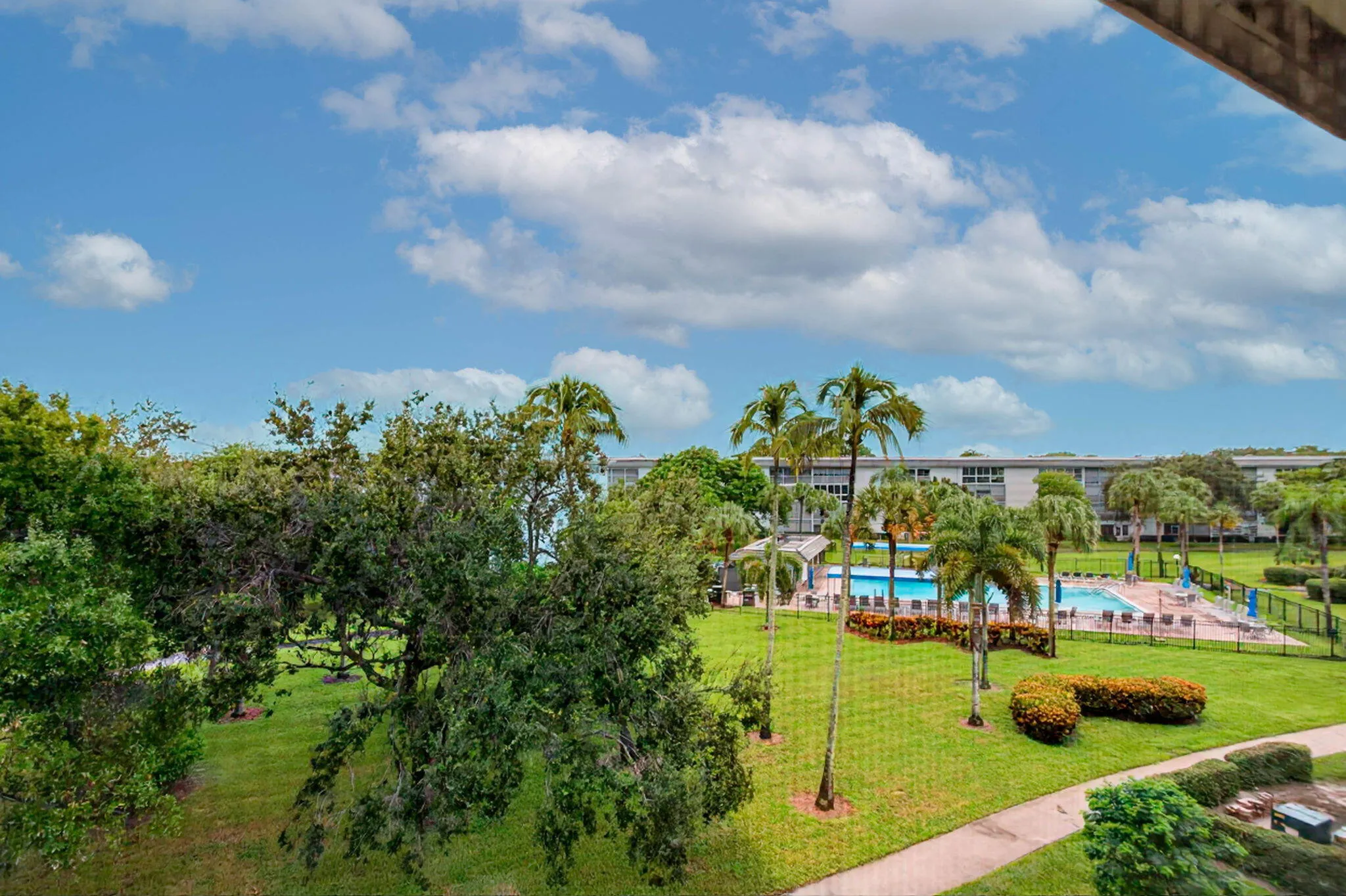 Expansive green spaces with mature trees and palms, featuring a fenced swimming pool with surrounding lounge areas