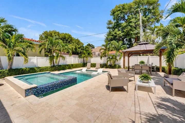 Resort-style backyard oasis featuring a resort-style pool with mosaic tile accents, expansive travertine patio, mature palm and shade trees, pergola-covered lounge area, and white perimeter fencing for privacy