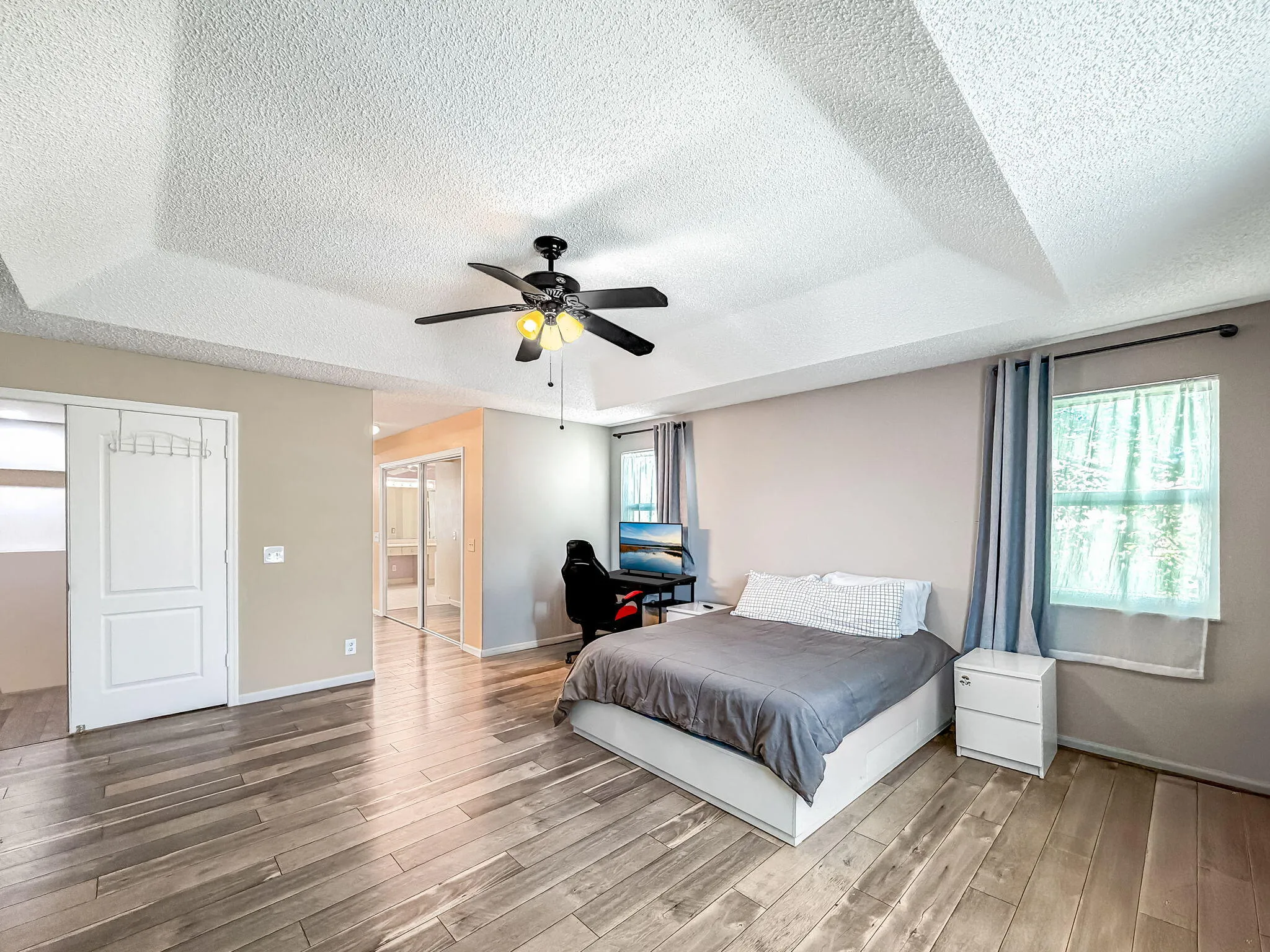 Tray ceiling with textured finish, wood-finish flooring, ceiling fan with integrated lighting, and multiple windows with curtain rods