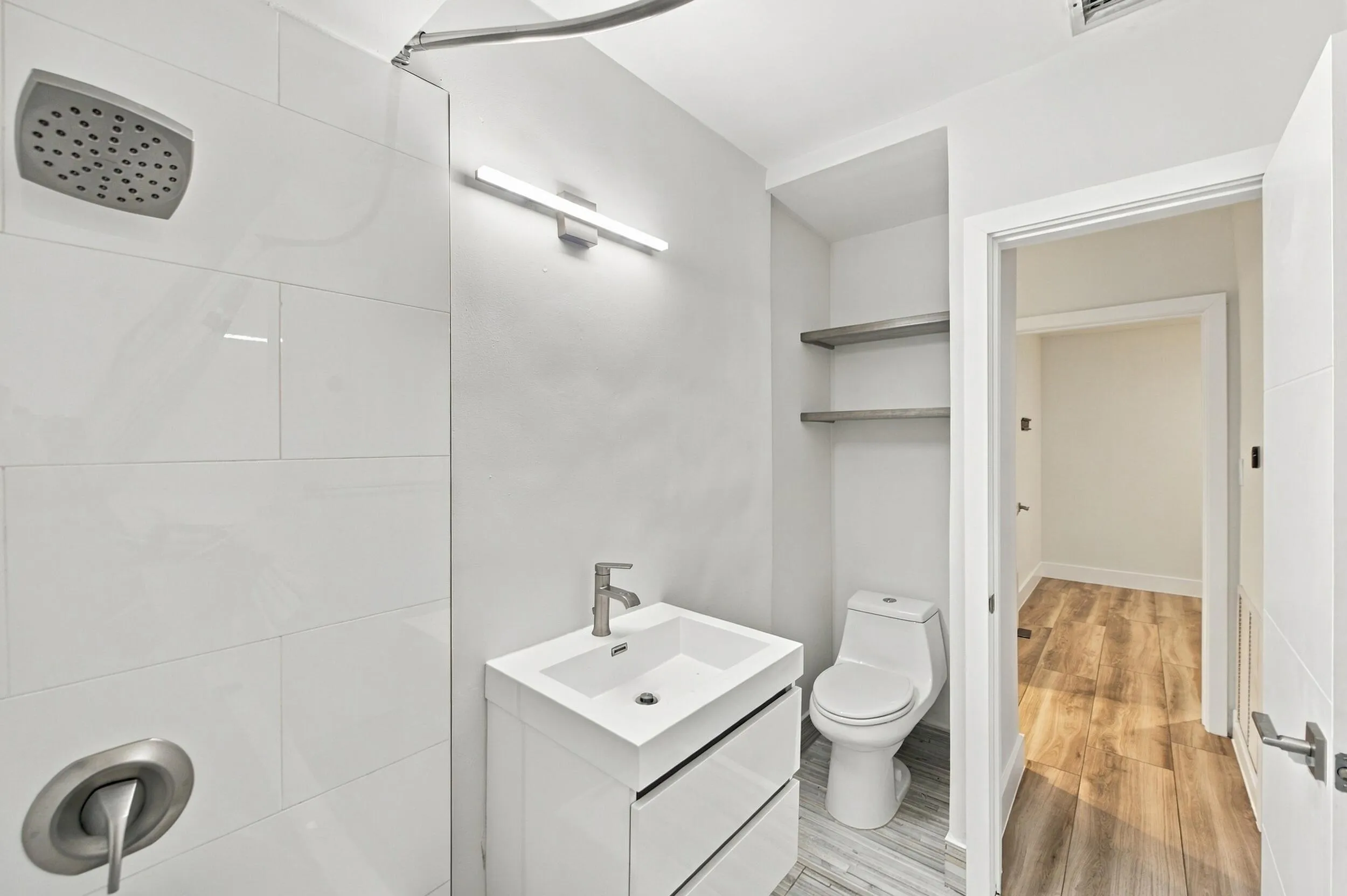 Walk-in shower with large format wall tiling, single vanity with integrated sink, modern wall-mounted light fixture, built-in shelving, and wood-finish flooring