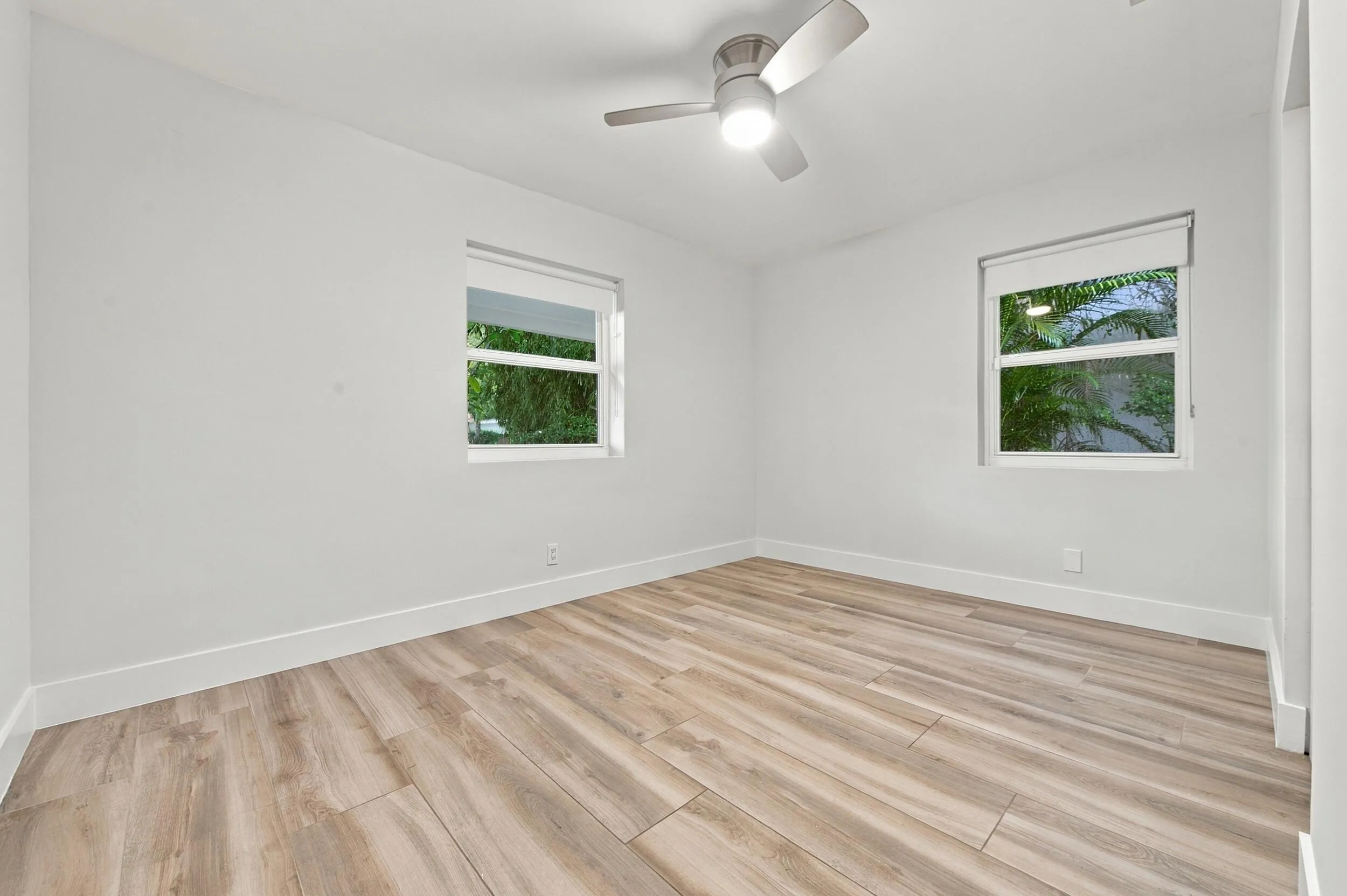 Light-toned wood-finish flooring, white walls, two windows with roller shades, and a ceiling fan with integrated lighting