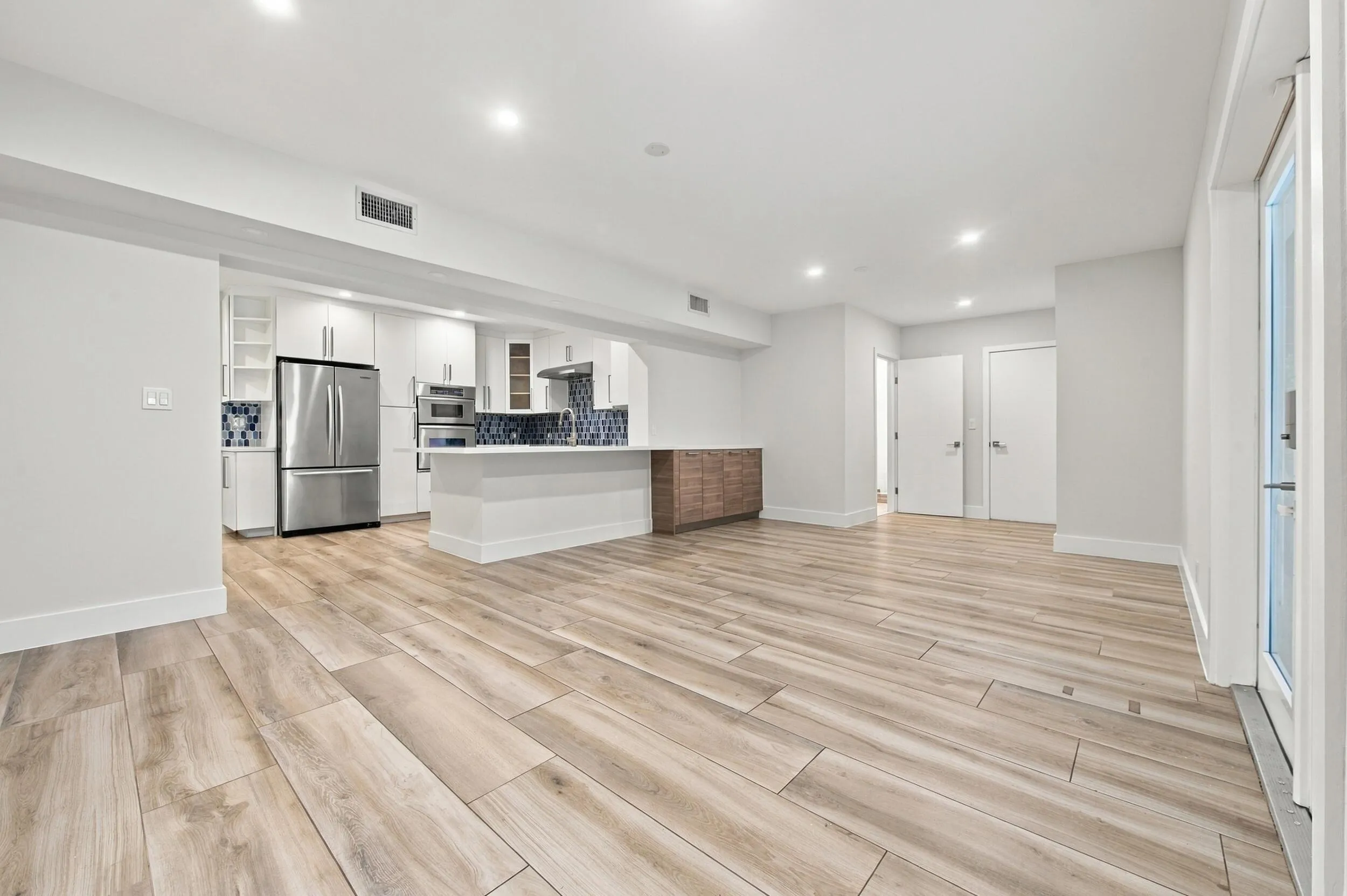 Open-concept living space with wood-finish flooring, recessed lighting, and a modern kitchen featuring stainless steel appliances, white cabinetry, and a blue tile backsplash