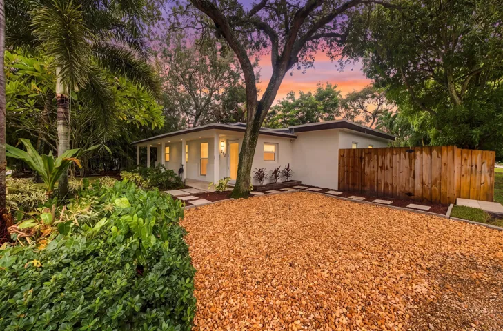White stucco exterior with a covered front porch, gravel driveway, wood privacy fence, and mature landscaping