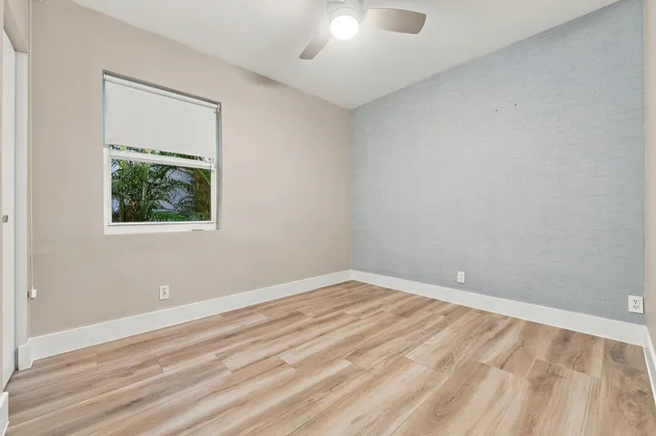 Wood-finish flooring, light grey accent wall, white trim, window with roller shade, and a ceiling fan with integrated lighting