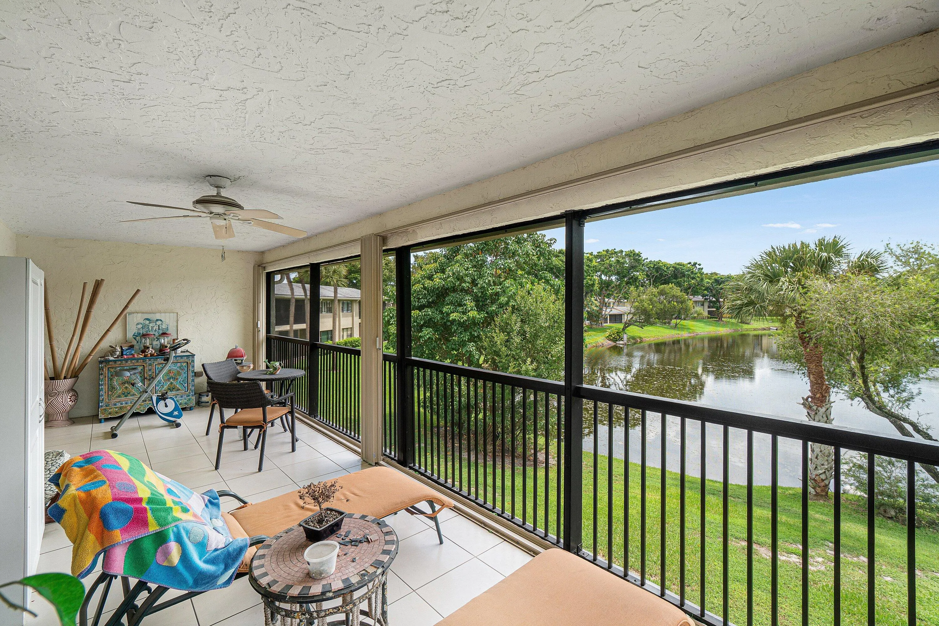 Screened balcony featuring tile flooring, a ceiling fan, and black metal railings overlooking a body of water with lush greenery