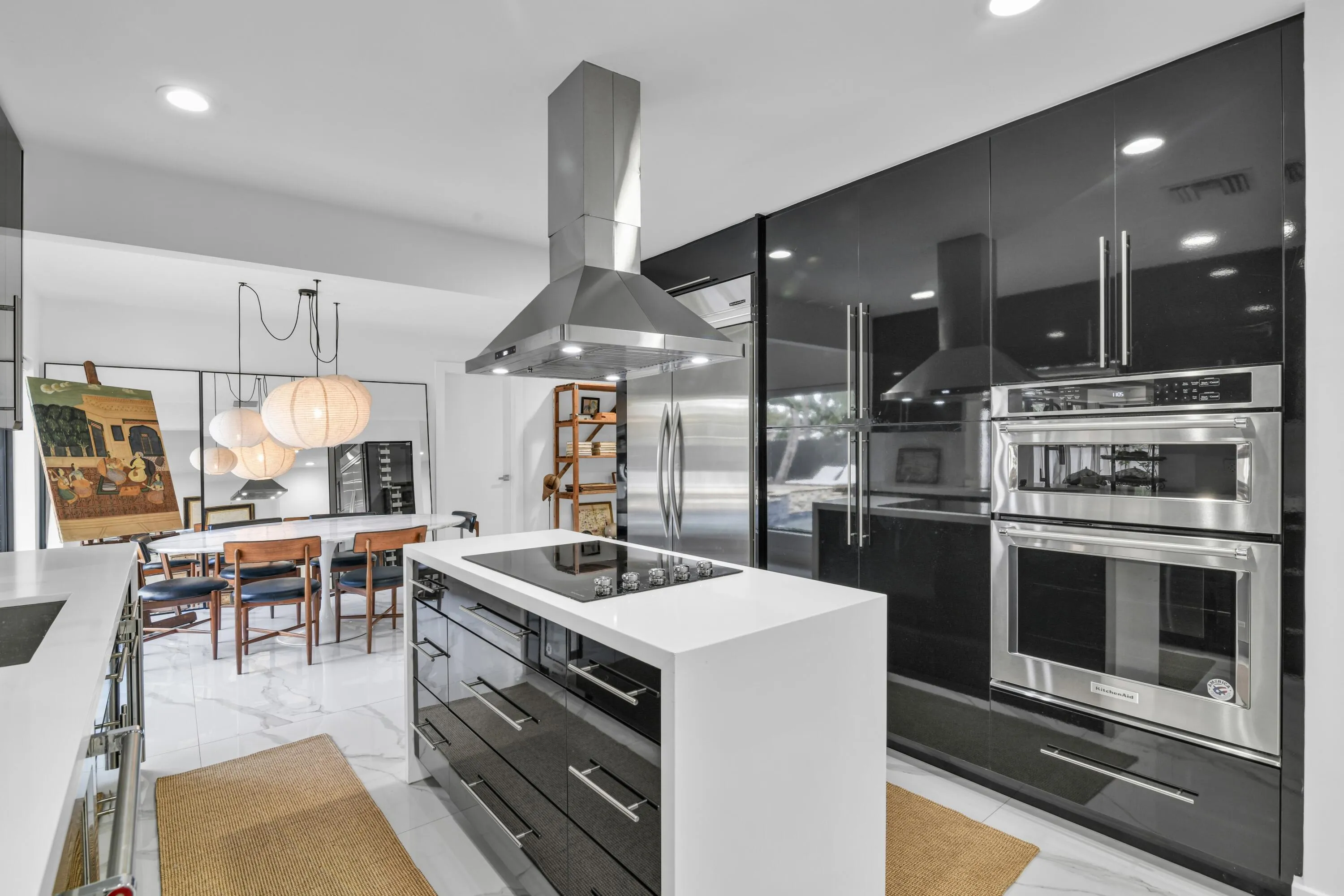 White countertop island with built-in cooktop, stainless steel range hood, high-gloss black cabinetry, stainless steel double wall oven, and recessed lighting