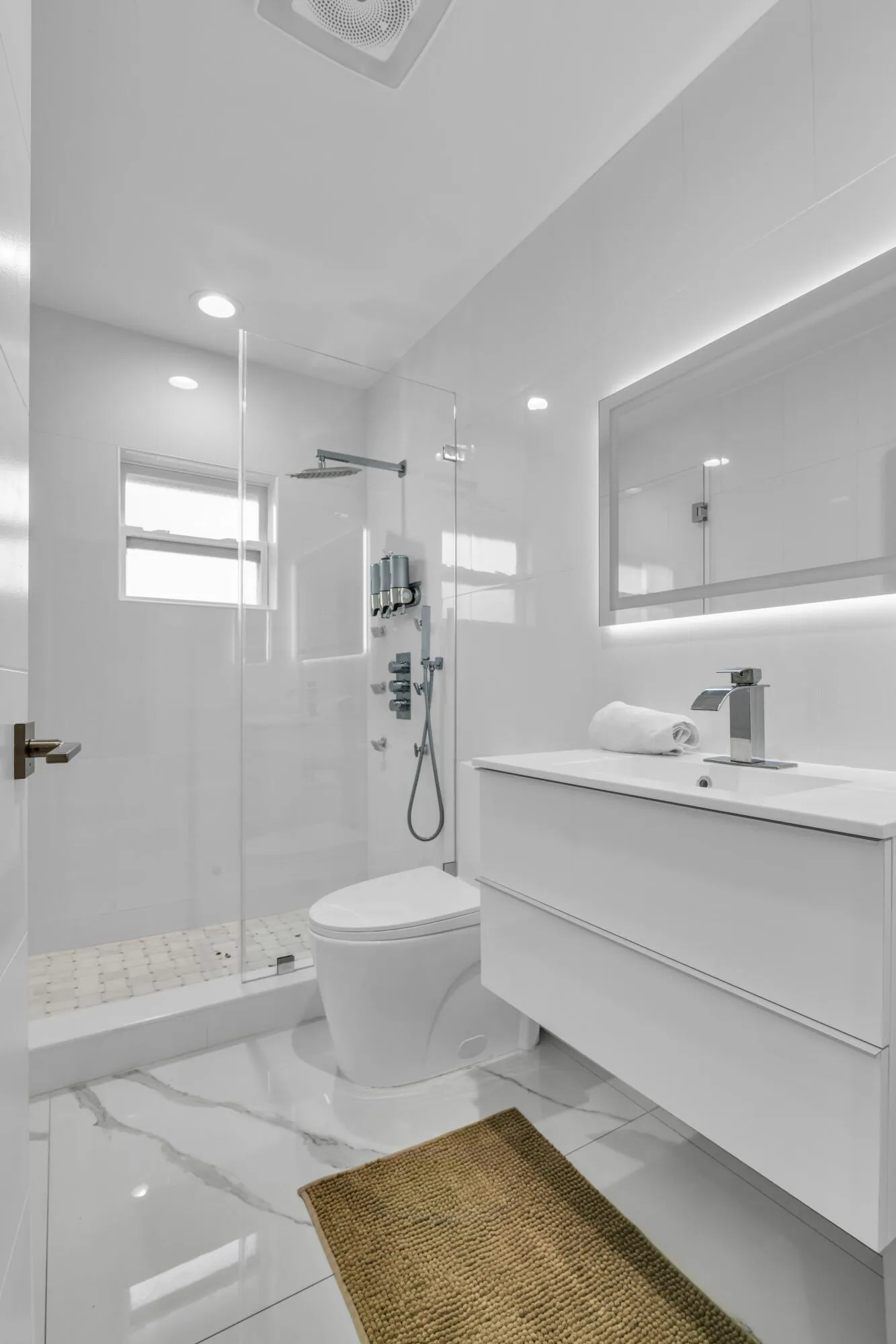 Floating vanity with integrated sink, glass-enclosed shower, wall-mounted shower system, illuminated mirror, and marble-look tile flooring