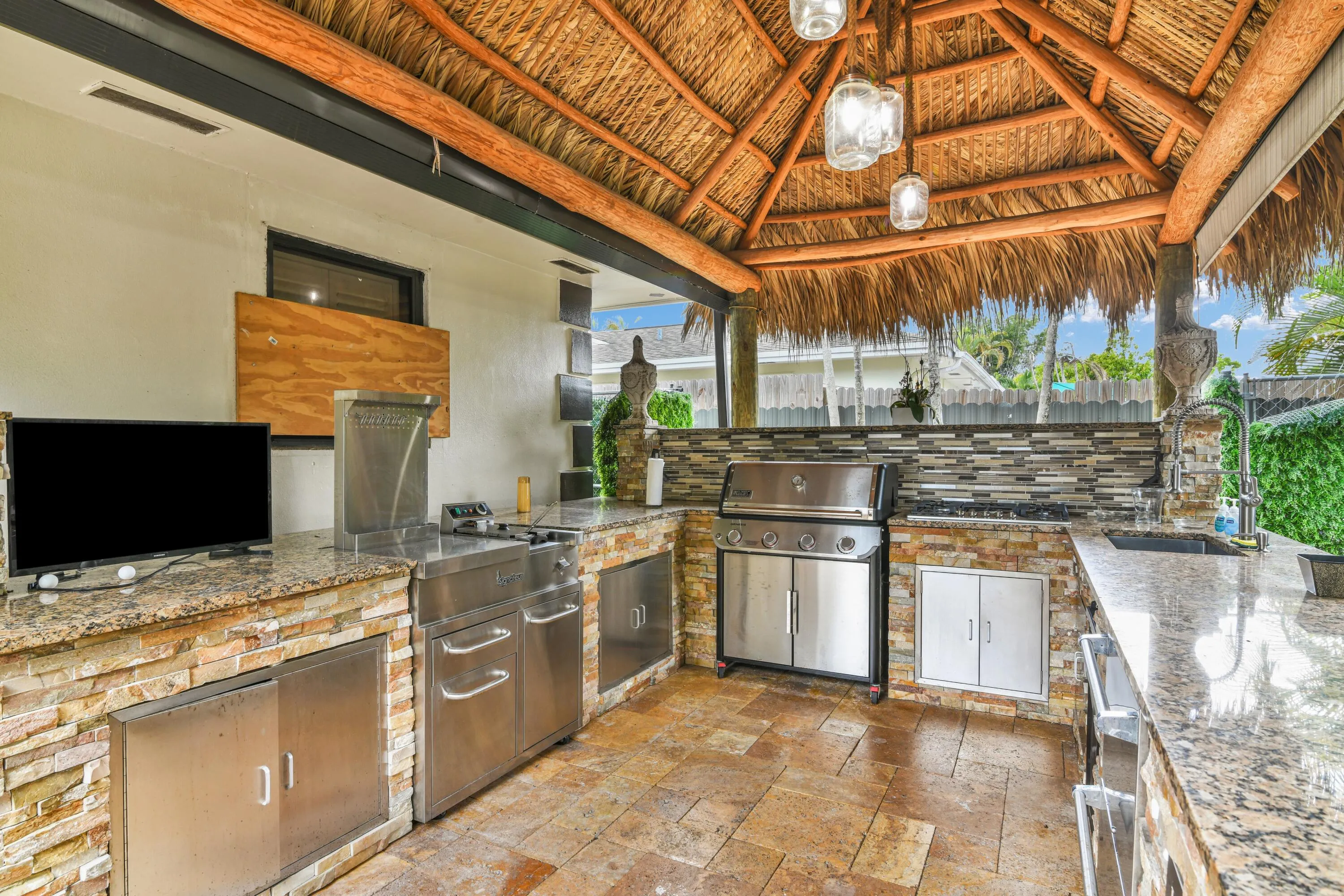 Outdoor kitchen featuring a thatched roof, stone-clad base, granite countertops, built-in stainless steel grill, and integrated sink