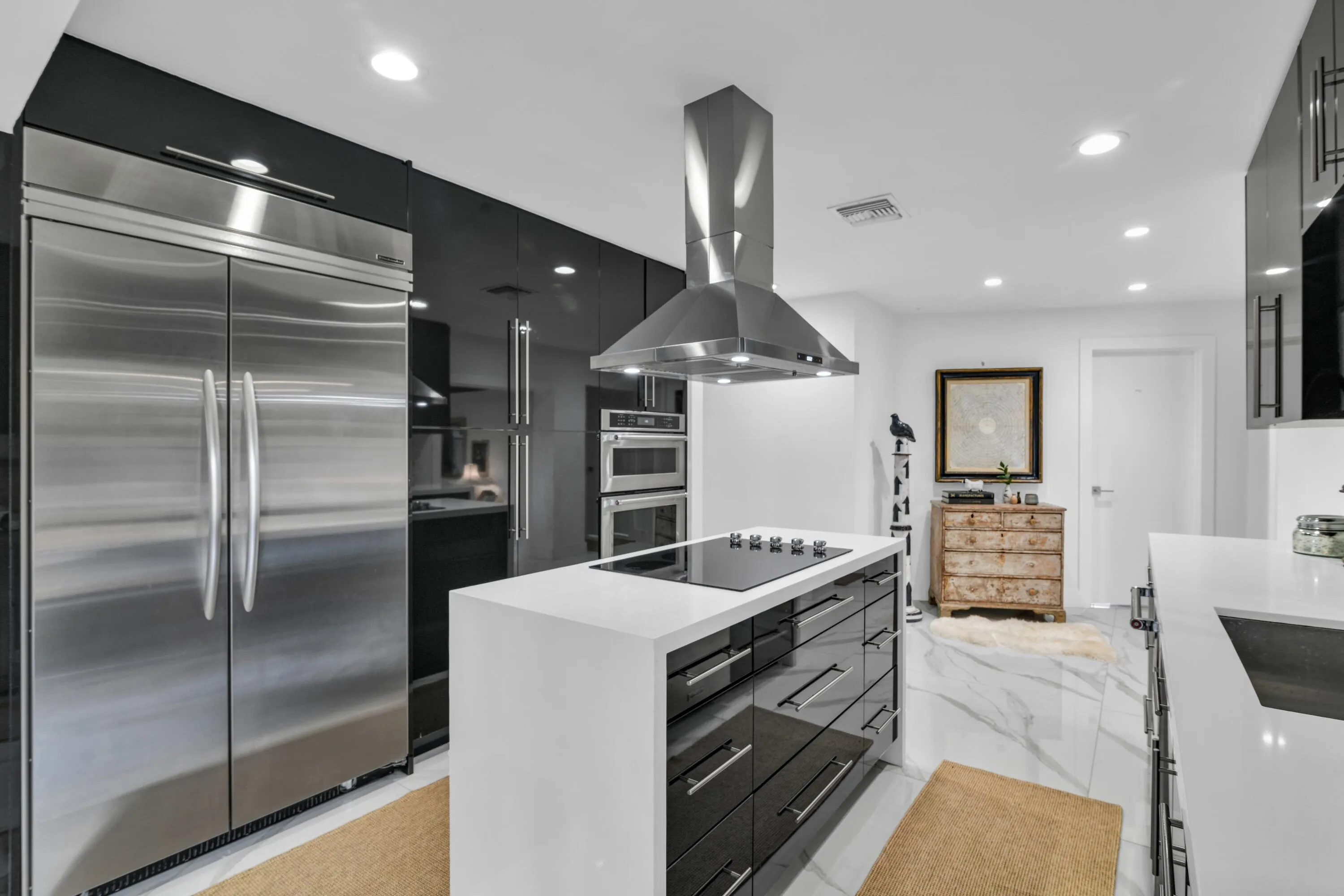 Stainless steel double-door refrigerator, sleek black cabinetry, stainless steel range hood, built-in double ovens, and a kitchen island with cooktop