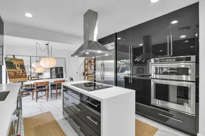 White countertop island with built-in cooktop, stainless steel range hood, high-gloss black cabinetry, stainless steel double wall oven, and recessed lighting