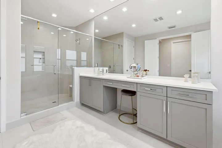 Spacious bathroom featuring a frameless glass shower enclosure, a full-width vanity mirror, light-toned tile flooring, recessed lighting, and gray shaker-style cabinetry with a built-in vanity area