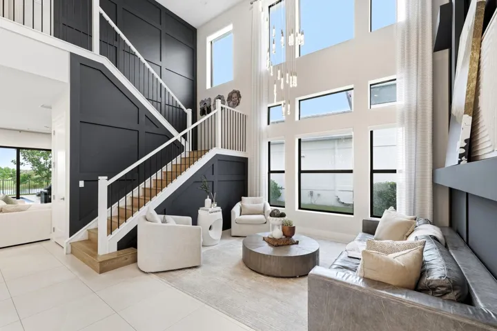 Two-story living area with high ceilings, multiple large windows, a built-in shelving unit, and a prominent staircase with wood-finish treads and an open balustrade