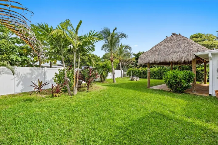 Expansive lawn featuring a tiki hut with thatched roof, mature palm trees, diverse tropical landscaping, and white privacy fencing