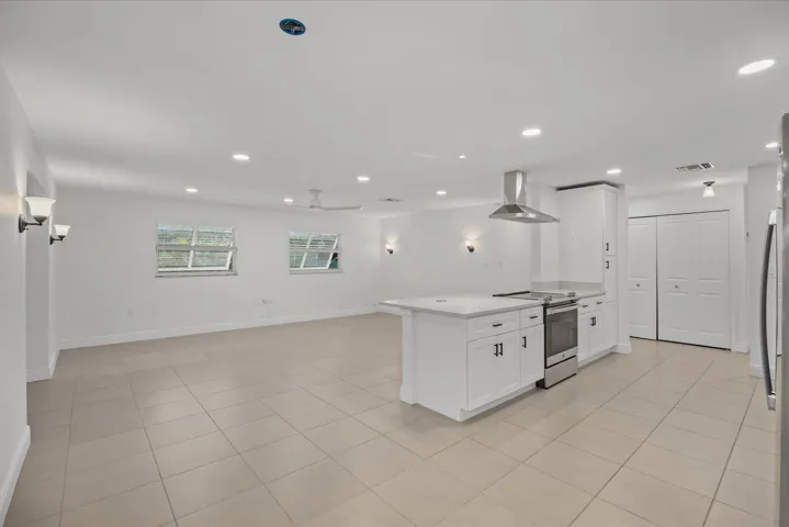 Spacious open-concept kitchen featuring a large center island with white cabinetry, stainless steel appliances including a dual-fuel range and range hood, neutral tile flooring throughout, recessed ceiling lighting, and abundant natural light from multiple windows