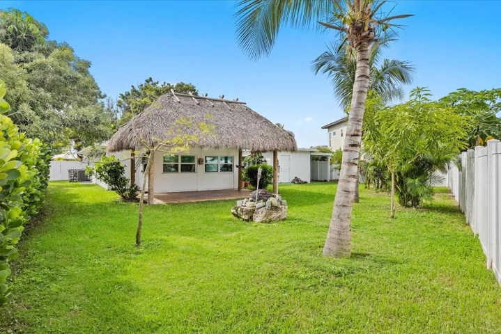 Expansive grassy yard featuring a thatched-roof structure, privacy fencing, and tropical landscaping