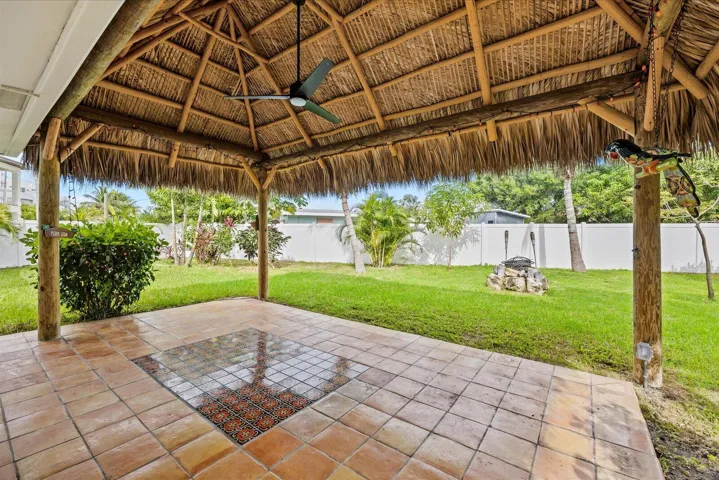 Covered outdoor entertaining space featuring a thatch-roof palapa structure with wooden support beams and ceiling fan, terra cotta tile flooring with decorative inlay detail, and expansive manicured lawn with mature landscaping and perimeter fencing
