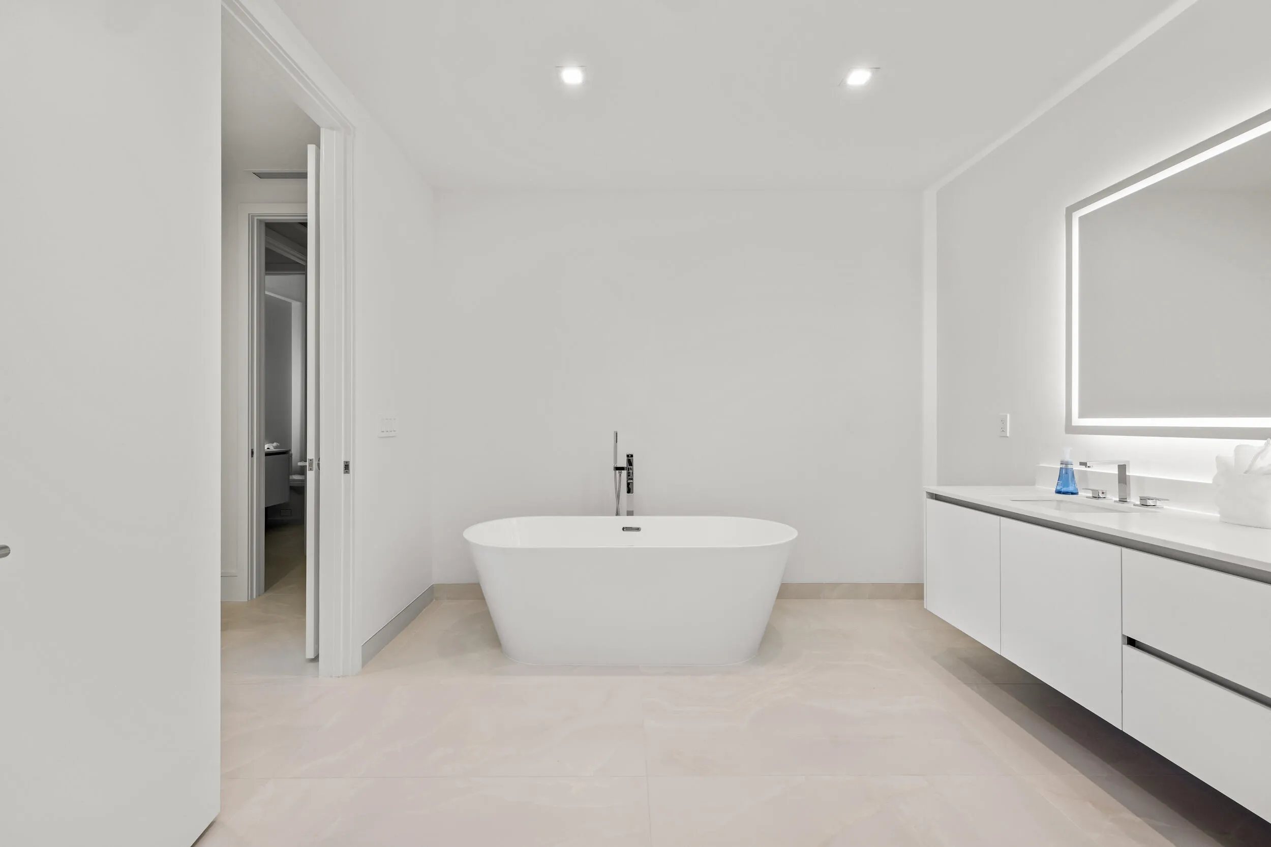 Master Bath - Freestanding soaking tub with floor-mounted faucet, dual-sink floating vanity with white flat-panel cabinetry, light-toned tile flooring, and an illuminated mirror