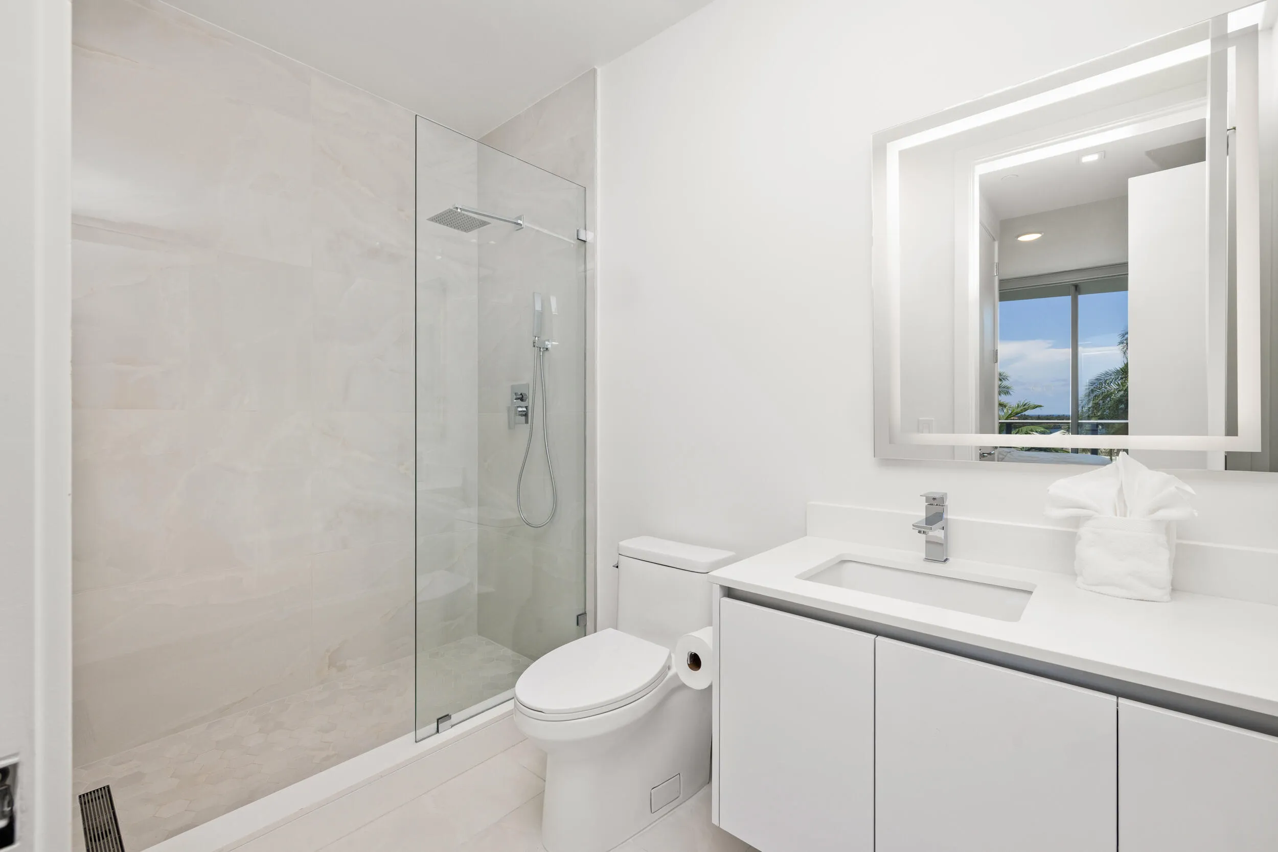 Contemporary bathroom featuring a frameless glass shower, rainfall showerhead, sleek vanity with integrated sink, chrome fixtures, and an illuminated mirror