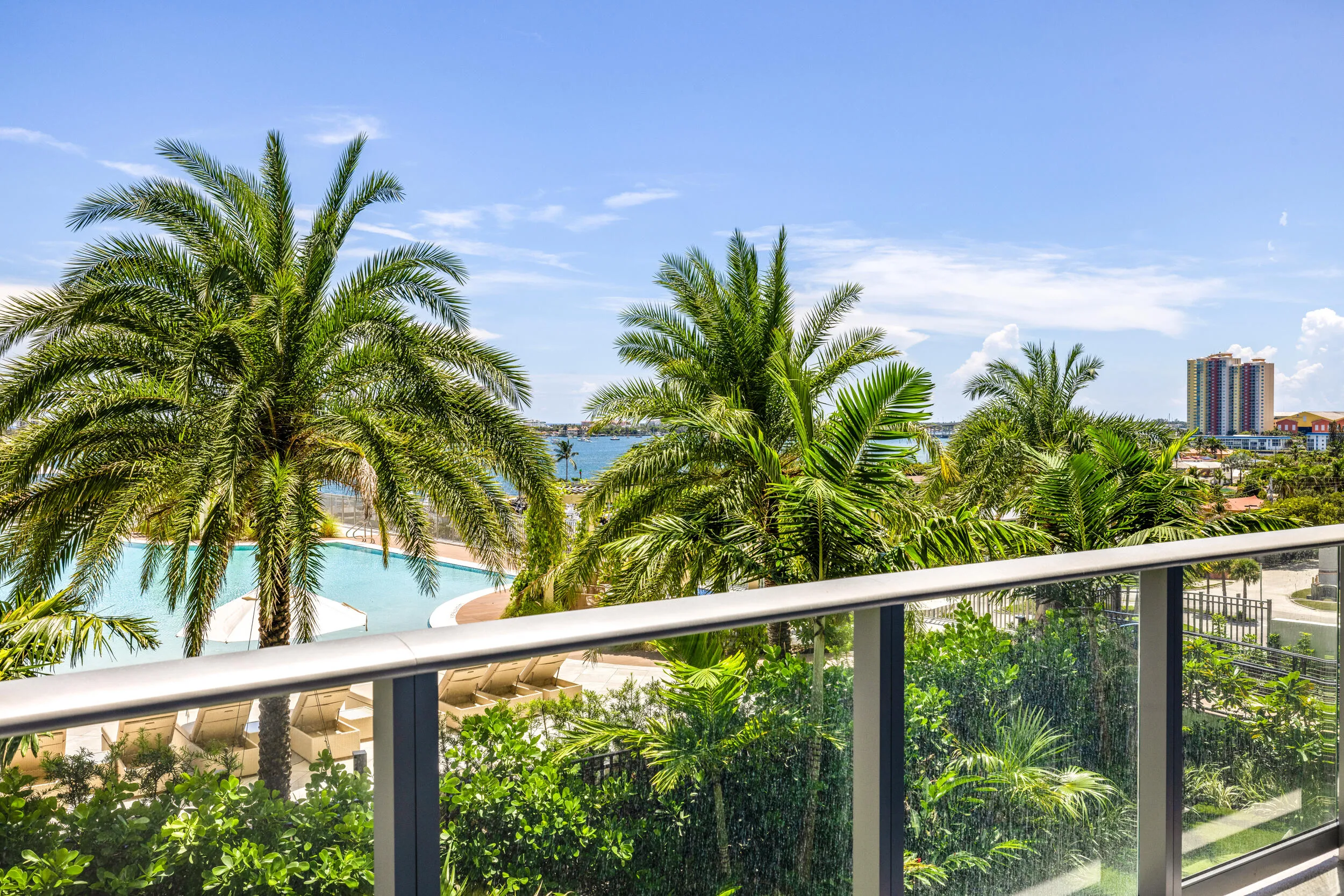 Glass balcony railing with aluminum supports, offering expansive views of tropical landscaping, a resort-style pool, and distant waterways