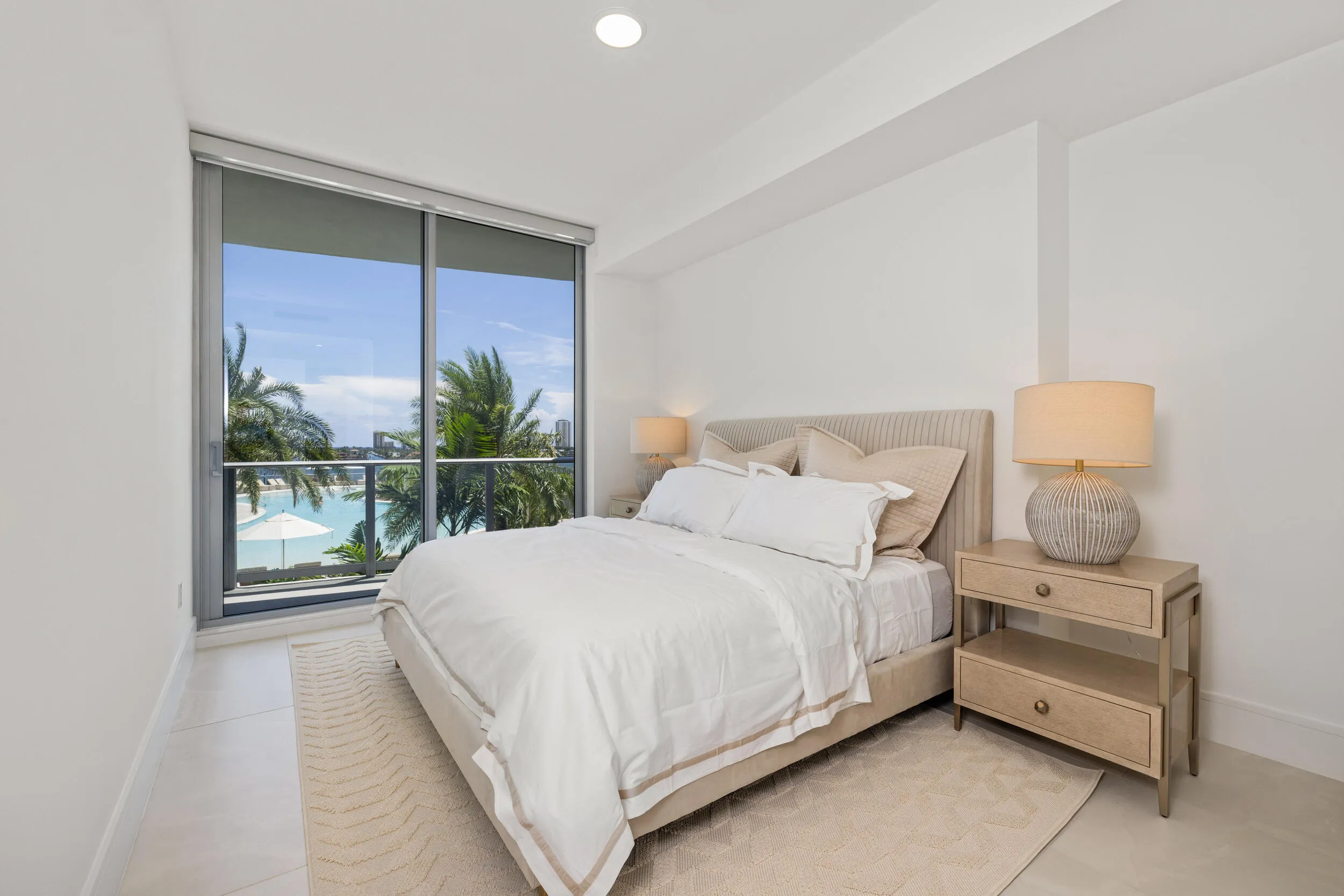Floor-to-ceiling glass doors, white tile flooring, recessed ceiling lighting, and a neutral upholstered headboard