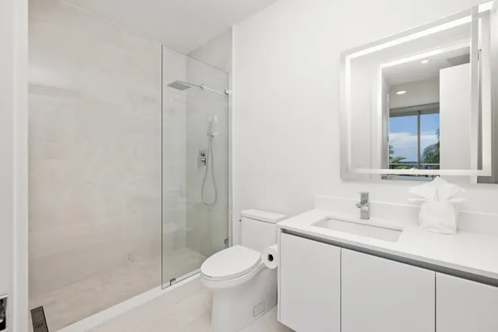 Contemporary bathroom featuring a frameless glass shower, rainfall showerhead, sleek vanity with integrated sink, chrome fixtures, and an illuminated mirror