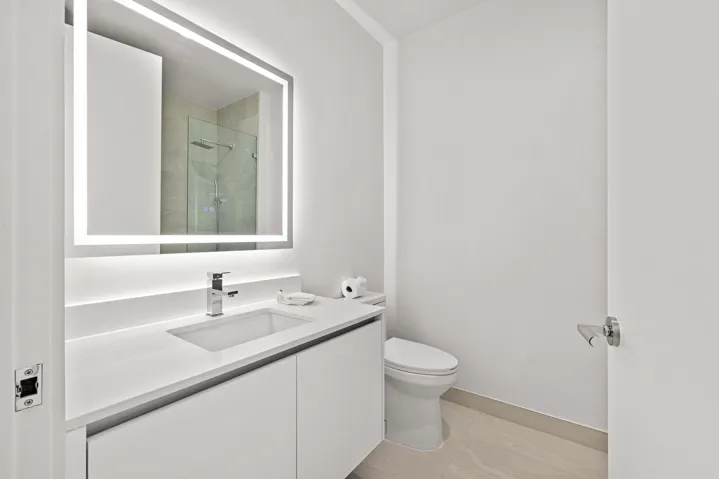 Backlit vanity mirror, single basin sink, white integrated countertop, white cabinetry, and a walk-in shower with glass enclosure