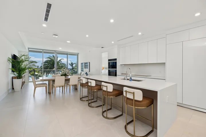 Sleek kitchen with white cabinetry and integrated appliances, expansive central island with countertop seating, open-concept living space, extensive floor-to-ceiling windows, and recessed lighting