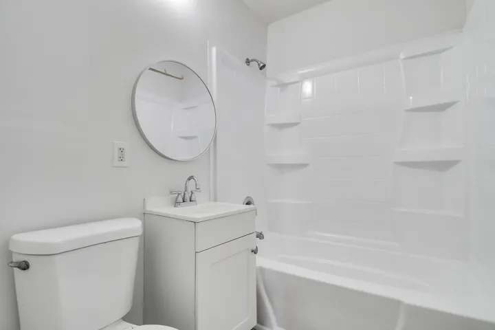 White vanity with chrome faucet, circular mirror, and combination shower/tub with integrated shelving