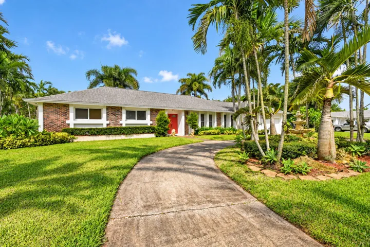 Brick facade residence featuring a red entry door, a curvilinear concrete driveway, mature palm trees, and a multi-tiered fountain
