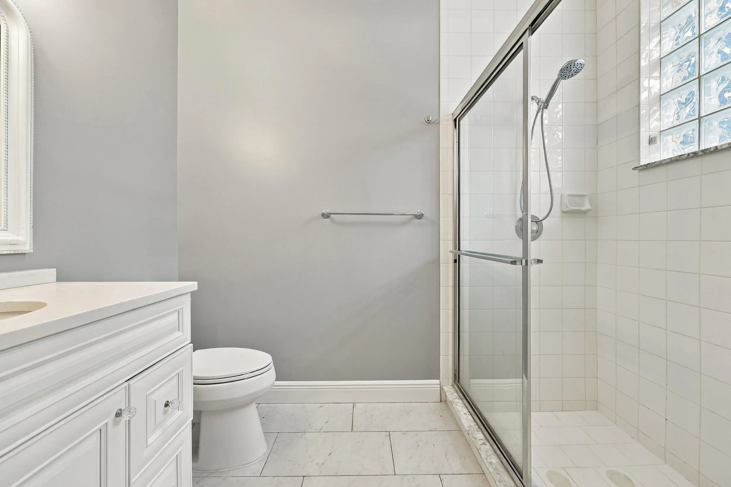 White vanity with clear knob hardware, white countertop, tile flooring, glass-enclosed shower, and block window