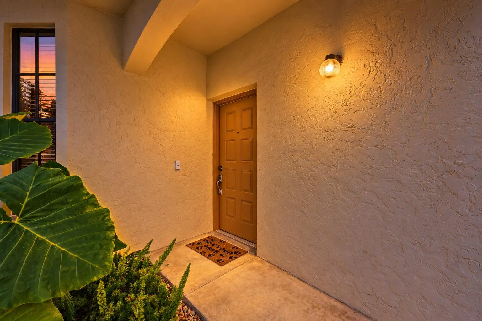 Textured stucco exterior with a paneled entry door, an exterior wall sconce, and a window with horizontal blinds