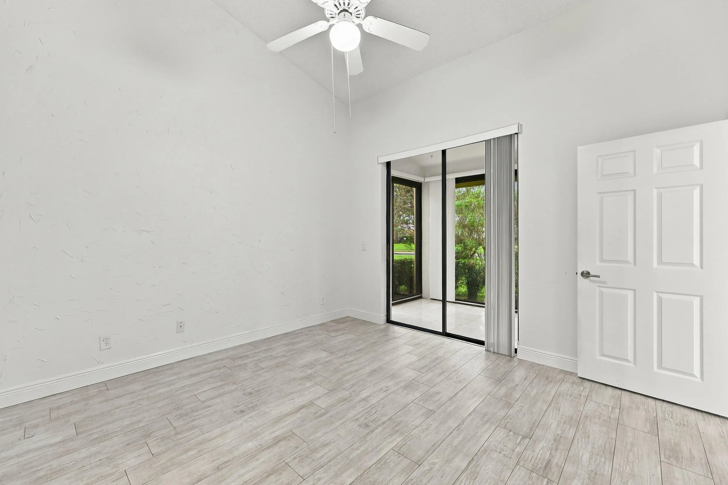 Vaulted ceiling with a white ceiling fan, sliding glass doors to an outdoor area, six-panel interior door, and light wood-finish flooring