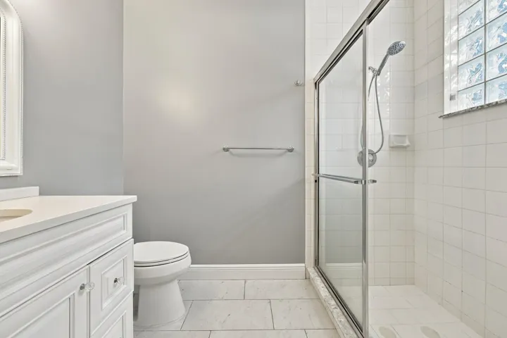 White vanity with clear knob hardware, white countertop, tile flooring, glass-enclosed shower, and block window