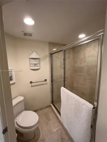 Recessed ceiling lighting, full-size toilet, tiled flooring, glass-enclosed shower, and a built-in towel bar