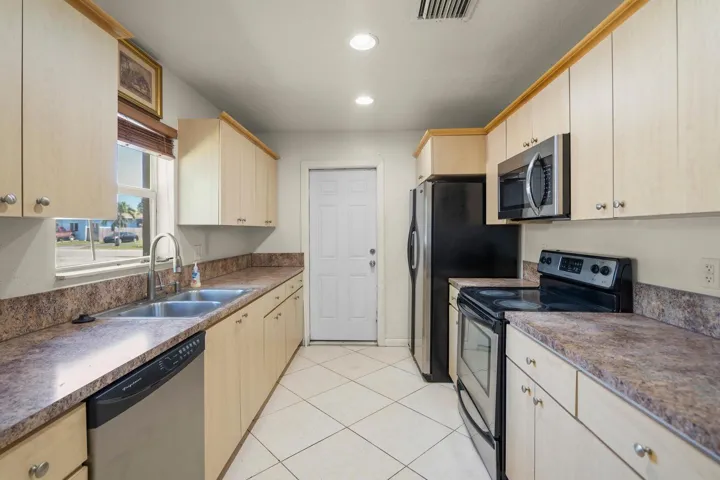 Galley kitchen featuring light wood-finish cabinetry, a double basin stainless steel sink with a gooseneck faucet, stainless steel dishwasher, and matching stainless steel microwave and range