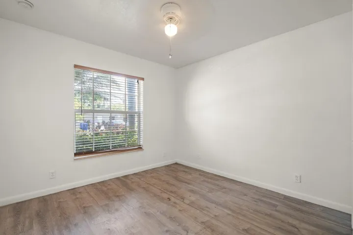 Wood-finish flooring with white baseboards, a window with horizontal blinds and a wood-toned frame, and a ceiling fan with integrated lighting