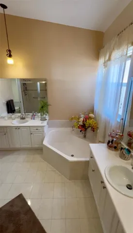 Bathroom featuring dual white vanities with integrated sinks, a built-in corner soaking tub, ceramic tile flooring, and a glass-enclosed shower