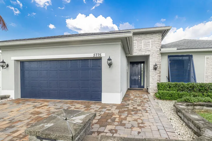 Stucco exterior with stone accents, paver driveway, dark blue garage door, dark blue entry door with decorative glass, and exterior sconce lighting
