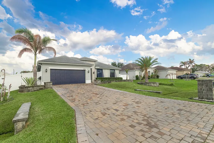Paver driveway, attached garage, well-maintained lawn, exterior lighting fixtures, and mature palm trees