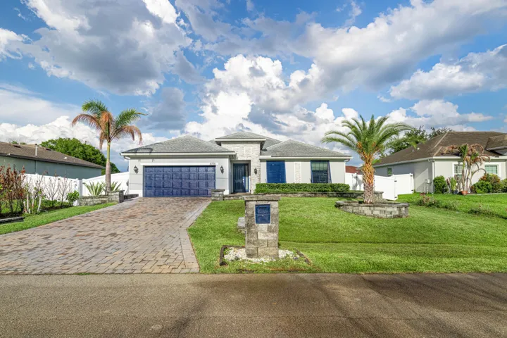 Contemporary single-story residence with a paved driveway, blue garage door, exterior wall sconces, and a lush front lawn with mature palm trees