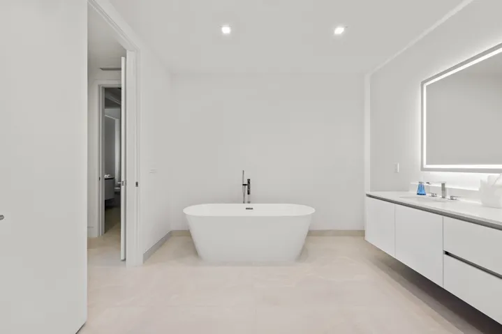 Master Bath - Freestanding soaking tub with floor-mounted faucet, dual-sink floating vanity with white flat-panel cabinetry, light-toned tile flooring, and an illuminated mirror