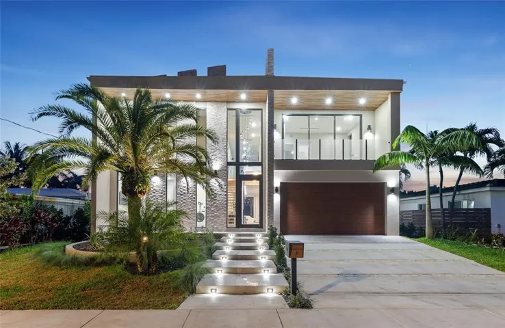 Modern exterior architecture featuring a prominent stone facade, floor-to-ceiling windows, a wood-finish garage door, a second-story balcony with glass railings, and illuminated exterior steps