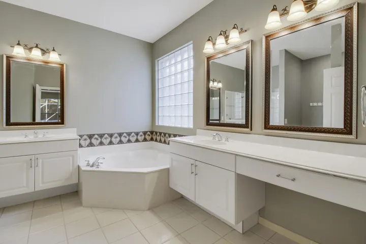 Dual vanity sinks with white cabinetry, a built-in soaking tub, decorative tile accents, and a glass block window
