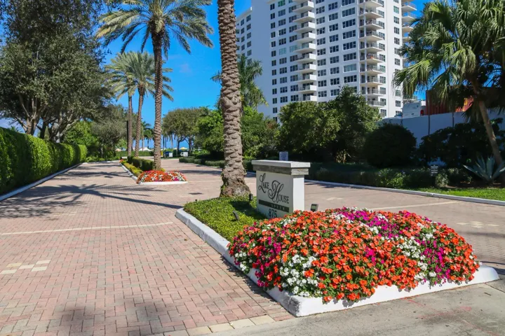 Paver driveway entrance with lush landscaping, mature palm trees, and a multi-story building exterior