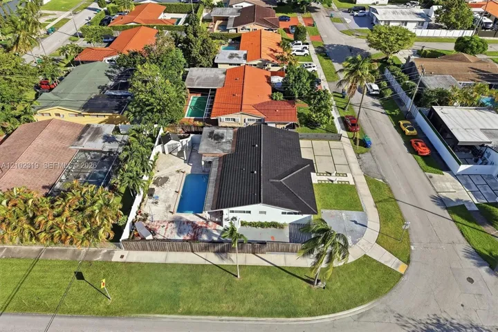 Aerial perspective of a corner lot property featuring a dark-toned roof, an in-ground swimming pool, a concrete-paver driveway, and fenced perimeter